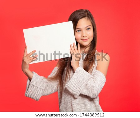 Beautiful European girl teenager 10 years old holds a white sheet of paper. On a red background. Place for text, banner, surface for inscriptions, advertising, text.