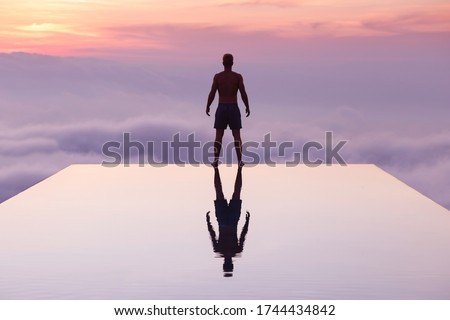 A young man standing on an infinity pool during sunset time. The pool was located high in elevation, so he felt like swimming into the clouds. Bali. Handsome young man. Fit. Reflection. Royalty-Free Stock Photo #1744434842