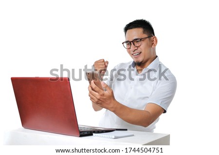 Portrait of excited Young Asian man sitting near laptop on the desk successfully completed the work as online freelancer and get payment, isolated on white