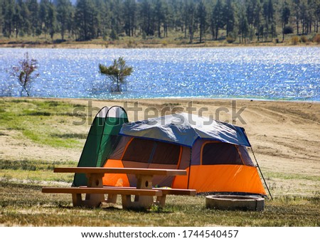 A camping tent and a pop up changing tent next to a picnic table and fire pit near the lake.