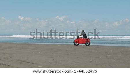 Handsome man biker surfer driving his black motorbike cafe racer with red surfboard shortboard on the beach along the ocean at sunny day