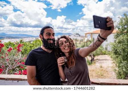 Couple of blonde curly hair woman and long black hair and beard man taking a selfie on a terrace with the town in the background