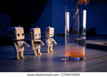 A small wooden toy robots looks at glass filled with soda in the dark. wallpaper, blured background. low light. concept of eco-friendly toys
