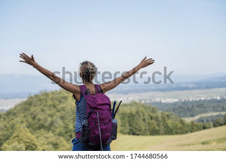 View from behind of a young female hiker with purple backpack on her back, standing and looking at beautiful view enjoying it with arms raised high.
