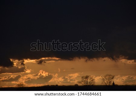 Monochromatic, golden colored landscape of a countryside with silhouettes of trees and a church tower framing the picture from the bottom and heavy rain clouds from the top. 