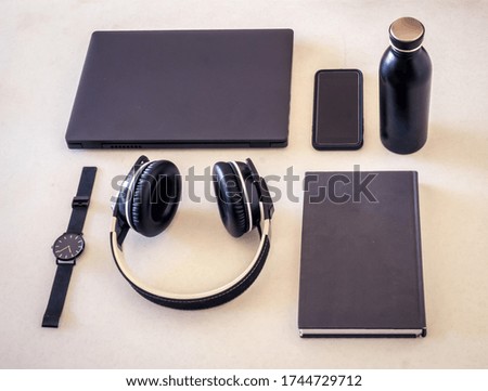 Organized black and grey accessories on a desk such as a laptop, a book, a watch, a phone a water bottle and a pair of headphones.