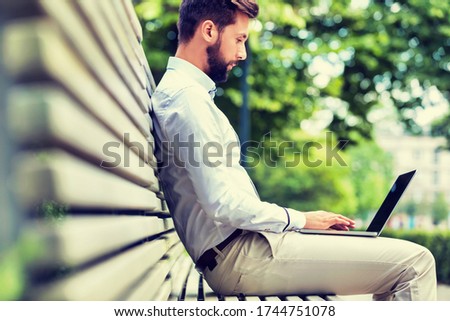 Portrait of young attractive businessman sitting on bench while working on his laptop
