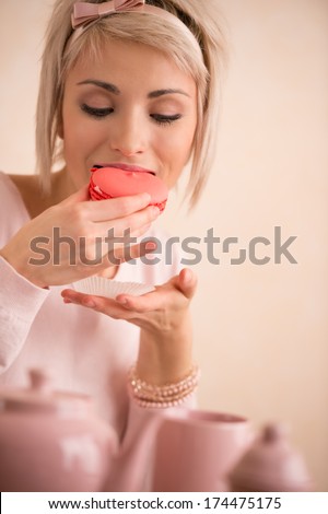 Young beautiful blond woman eating macaroon while having tea-party. She is very satisfied. Short hair and pink colors - modern style