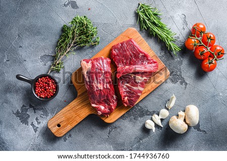 Raw flap steak flank cut with Machete, Skirt Steak, on woods chopping board, with herbs tomatoes peppercorns over grey stone surface background top view Royalty-Free Stock Photo #1744936760