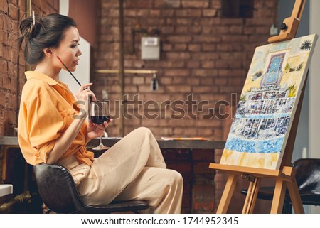 Beautiful female artist looking at abstract painting and holding glass of wine while sitting on stool in front of easel