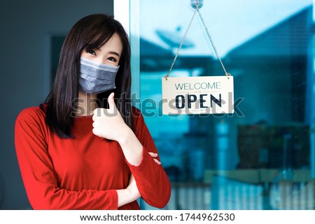 Asian woman shop owner smiling wearing medical face mask hanging label 'Welcom we are open' and showing thump up. She open her shop after pandemic of coronavirus outbreak