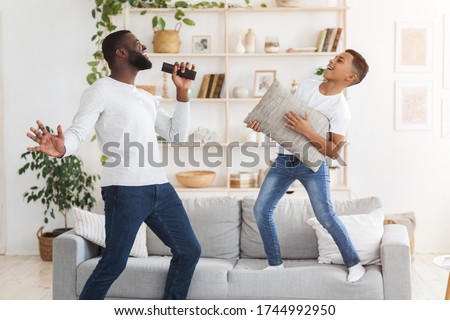 Home Entertainment. Cheerful black dad and preteen son having fun together in living room, singing and dancing, boy using pillow as guitar