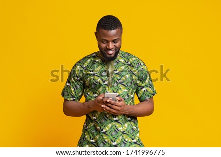Cheerful young african man looking at smartphone screen over yellow background, chatting with friends
