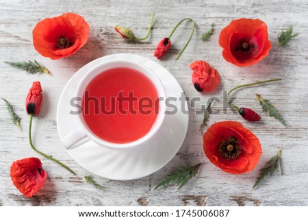 Berry tea in the white ceramic cup with saucer, poppy flowers,  buds and leaves on the rustic wooden background. Flat lay