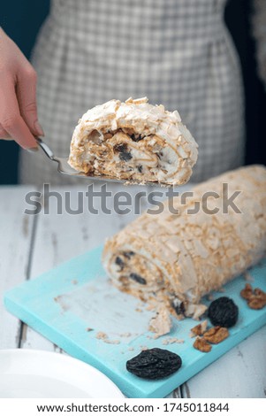 Woman serves with cake server a piece of meringue roll on a white vintage wooden kitchen table. Meringue pie decorated with prunes and walnuts on blue cutting board.