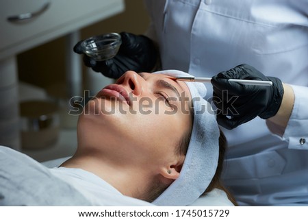 A female cosmetologist wearing black disposable medical gloves uses a brush to apply a superficial transparent face peeling to a woman. A cosmetology procedure in a beauty salon for skin cleaning. Royalty-Free Stock Photo #1745015729