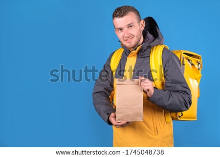 Young smiling caucasian food delivery man in yellow uniform and cooler bag on his back holds paper craft bag with ready order for customer on blue studio background. Courier delivers products home