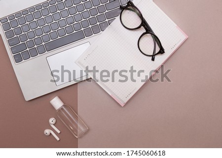Home office in quarantine. Covid-19. Glasses, an open notepad lying on top of a laptop,sanitizer,and headphones on a two-tone beige background.