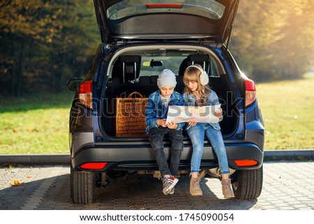 Agreeable boy and girl are looking at the road map while sitting in the auto's trunk and discussing the move direction. Family vacation trip by car. Royalty-Free Stock Photo #1745090054