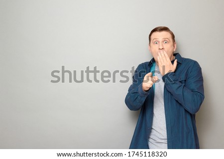Studio portrait of shocked frightened man wearing shirt, looking with eyes full of fear and pointing with finger at something awful, covering mouth with hand to prevent screaming, copy space on left