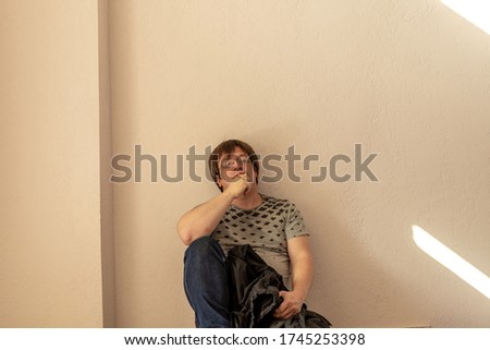 A man with glasses wistfully sits leaning on a wall. He's wearing jeans and a t-shirt. In his hand he holds a raincoat. High quality photo