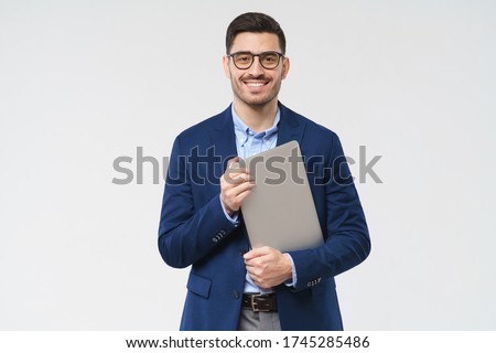 Young modern male teacher holding closed laptop in hands, waiting for students, smiling confidently, isolated on gray background