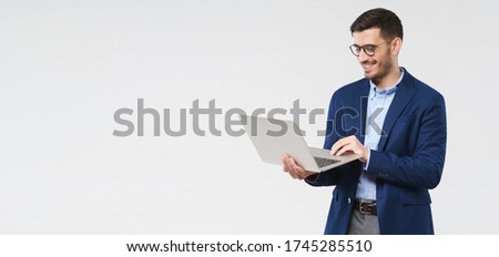 Horizontal banner of young handsome smiling business man holding laptop in hands, typing and browsing web pages, isolated on gray background with copy space