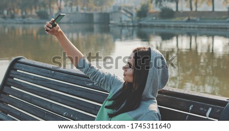 Brunette hipster woman wearing in hoodie selfie in park . Caucasian girl having fun with mobile phone , texting sending and reading messages , making photos