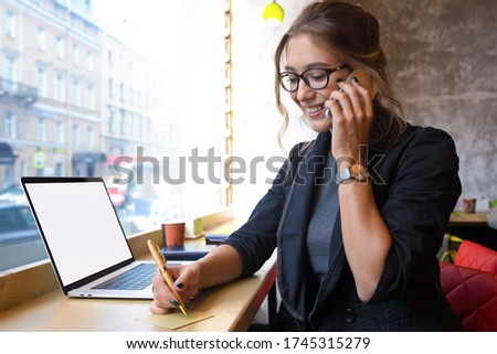 Cheerful woman in glasses clever University student talking via cellphone during online learning on laptop computer with empty mockup copy space display background for advertising 