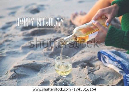 A girl sits on the beach and pours wine into a glass, a picnic near the sea. Lettering happy birthday.