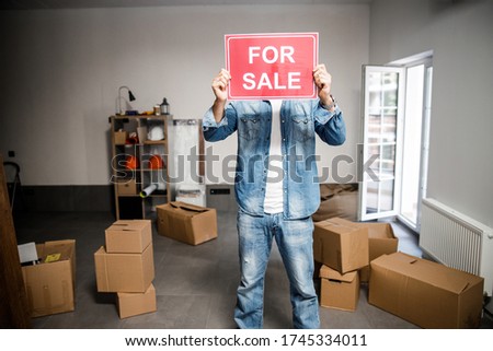 Male in denim wear is standing inside room with carton boxes and closing face with advertisement about selling apartment