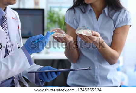 Patient holds breast implants next to doctor. Visit to female breast plastic surgery clinic. Consultation with an experienced surgeon. Support for women with cancer. Female breast implant selection Royalty-Free Stock Photo #1745355524
