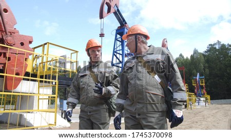 Two man oil workers walking and talking near oil pump jacks. Engineers overseeing site of crude oil production. Balancing drive rod pumps oil rocker. Equipment for wellhead connection well.