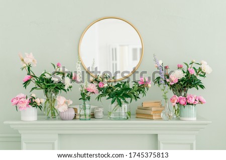 Pink and white peonies on the bookshelf  in glass vases. Round golden mirror on the wall above the fireplace