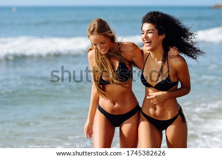 Two young women with beautiful bodies in swimwear on a tropical beach. Funny caucasian and arabic females wearing black bikini walking along the shore.