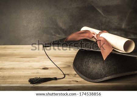 A diploma scroll tied with red ribbon, resting on a graduation mortarboard, on top of a wooden desk in front of a school blackboard.  Created in vintage style with low saturation. Royalty-Free Stock Photo #174538856