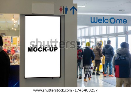 Mock up blank vertical LED board with clipping path in black frame on wall of shopping mall, blurred people wearing winter coat, empty space for insert advertising or announcement information