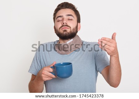 Picture of charismatic good looking young man closing eyes, holding cup with hot drink, enjoying smell of coffee, wearing pajamas, spending time in peace in morning alone. People and coffee concept.