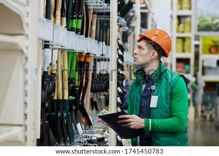 handsome guy warehouse worker counts quantity of shovels in store, make notes, wearing protective helmet and green uniform