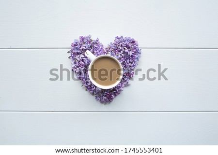 Heart shaped lilac flowers and a mug of hot coffee with milk on a white wooden background. The concept of good morning and the day of all lovers. Top view, flat layer, copy space for text.