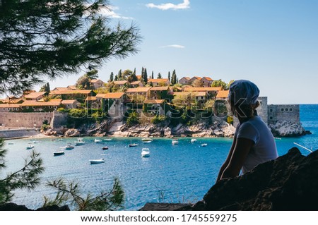 A girl in a white polka-dot panama admires the view of the island of St. Stephen in Montenegro. Sunny view of
island among green thickets is surrounded by a blue lagoon with white small boats.