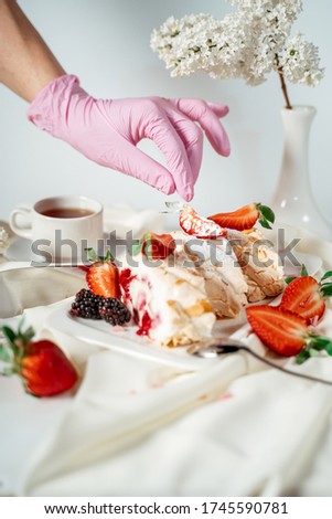 A gloved hand decorates a meringue cake, garnished with nut chips and strawberries, among lilac flowers. Food photography. Advertising and commercial close up design