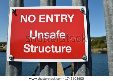 Isolated red and white rectangular no entry unsafe structure sign on metal posts with blurred harbour background on sunny day with blue sky