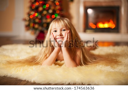 adorable girl with long hair lies on the fur near the fireplace and a Christmas tree 