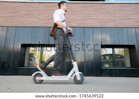 Blurry young businessman on scooter moving forwards along wall of contemporary business center while going home after work Royalty-Free Stock Photo #1745639522