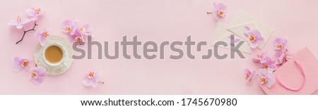 Delicate flatlay composition with morning cup of coffee with milk or cappuccino, letters, pink gift bag and purple orchid flowers on light pink background. Beautiful breakfast concept. Long banner