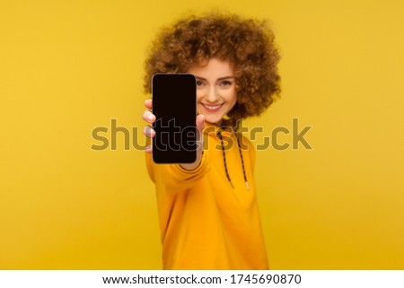 Portrait of cheerful curly-haired woman with engaging smile showing cellphone with empty display, mock up for app advertise, mobile web service. indoor studio shot isolated on yellow background