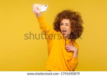Portrait of carefree trendy curly-haired girl in urban style hoodie taking selfie and grimacing with tongue out, having fun while doing photo, vlog broadcast. studio shot isolated on yellow background