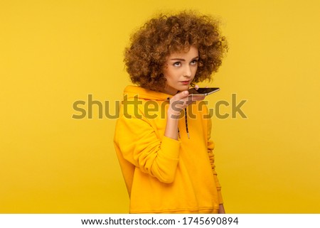 Virtual voice assistant on smartphone. Portrait of curly-haired woman in urban style hoodie giving command to mobile device in her hand, recording message. studio shot isolated on yellow background
