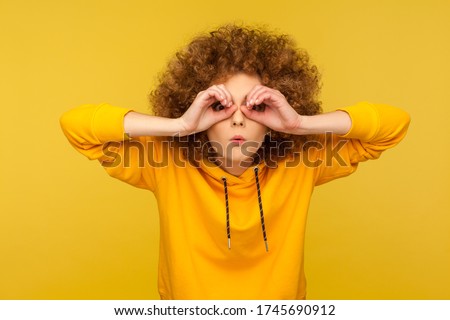 What is there? Portrait of surprised curly-haired woman in urban style hoodie making glasses shape, looking through binoculars gesture with shocked expression. indoor studio shot, yellow background